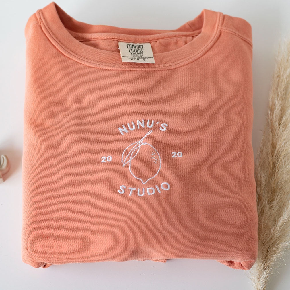 a salmon colored sweatshirt with a white logo embroidered on it boho staged