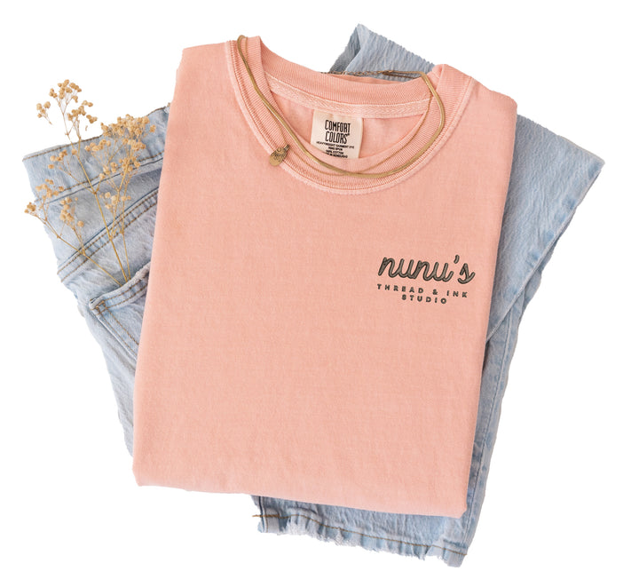 A peach t-shirt folded with jeans that is featuring a custom embroidered logo