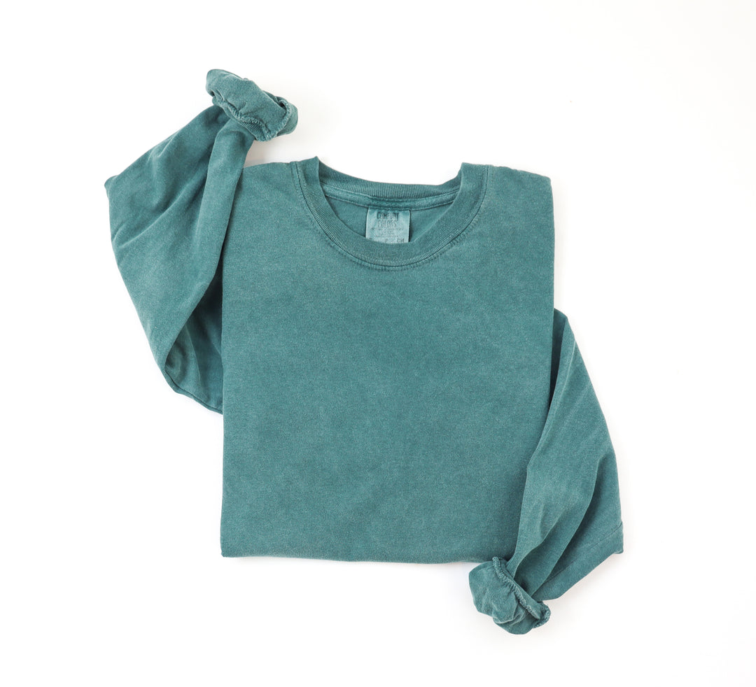 A neatly folded blue spruce colored long sleeved t-shirt on a white surface